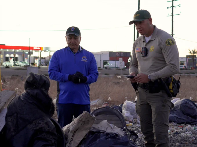 Two individuals, one in a San Bernardino County Sheriff’s uniform and another in a royal blue jacket, stand outside and engage with a homeless individual.