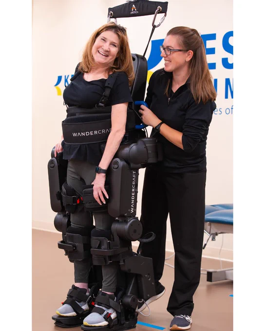 Image of a woman in a Wandercraft exoskeleton. There is a physical therapist next to her, smiling and assisting. 