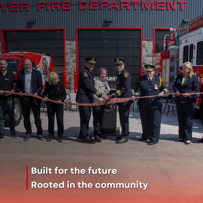 A group of people, including members of the Hanover Fire Department, participate in a ribbon-cutting ceremony outside a fire station. Red fire trucks are visible in the background.