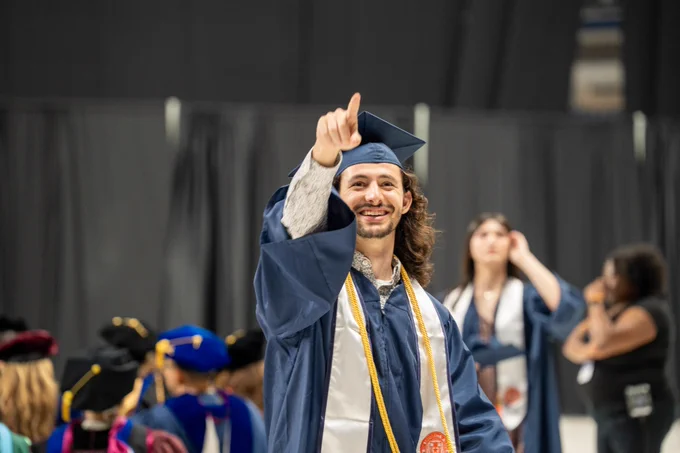 A graduate in a blue cap and gown makes a pointing gesture while smiling at a graduation ceremony.