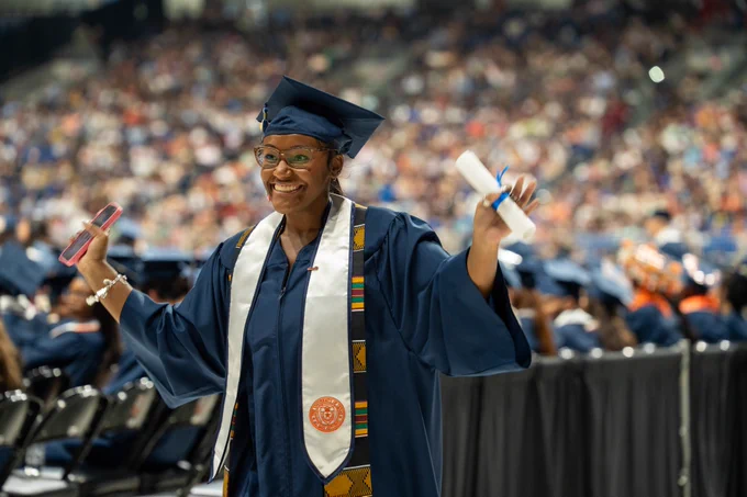 Graduate in cap and gown holding a diploma and phone, smiling at a university graduation ceremony.