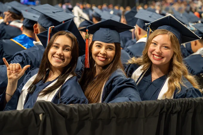 Three graduates in blue caps and gowns smiling at a graduation ceremony.