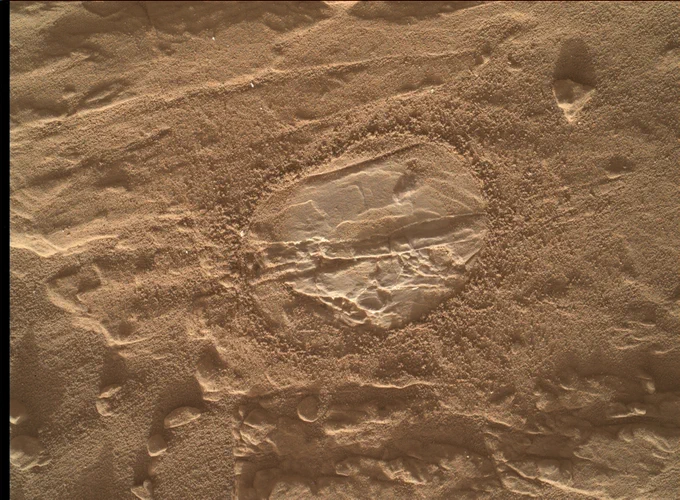 A close-up color view shows an oval shape where the Curiosity rover's dust removal tool swiped away Martian grit. The beige-colored surface is smooth and shows ridges across the rock. Surrounding the clean spot is thick Martian dust along a pockmarked surface.