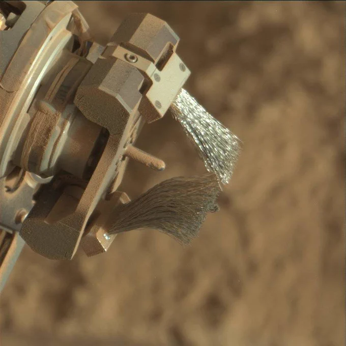 A close-up view shows the Curiosity rover's Dust Removal Tool (DRT) on Mars. The image shows two metallic wire brushes extending from a rotating mechanism, lightly coated with Martian dust. The blurred background reveals the reddish, sandy Martian surface.