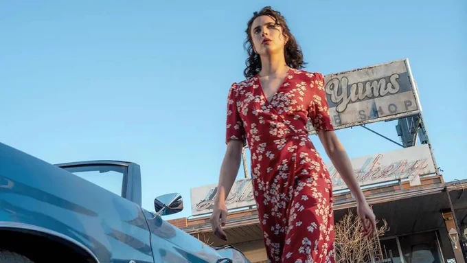 Margaret Qualley walks confidently alongside a car in a red dress in a still from Ethan Coen and Tricia Cooke’s new film HONEY DON'T!
