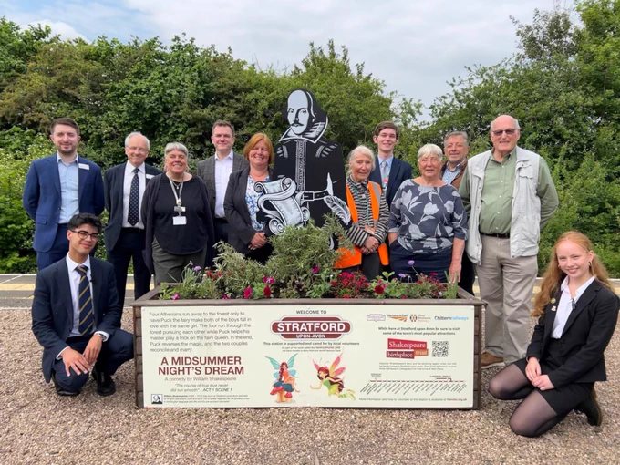 A group of 12 people, including what appear to be railway staff, community volunteers and students, pose on a train platform around a decorative planter. The planter features a large black silhouette of William Shakespeare, along with signage. The sign on the left of the planter has text that reads "A MIDSUMMER NIGHT'S DREAM" and describes the play's plot. The sign on the right welcomes visitors to Stratford-upon-Avon, mentions the Shakespeare birthplace and includes a QR code. The platform has green foliage in the background.