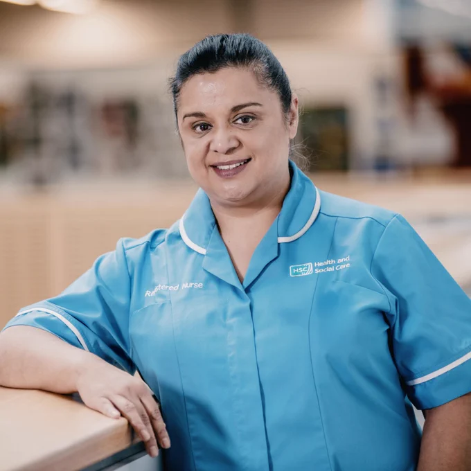
A female healthcare professional wearing a teal blue uniform with white trim, a name badge, and the logo "HSC Health and Social Care" on the right side of her chest, leans on a light-colored railing and smiles at the camera. She has dark hair pulled back.