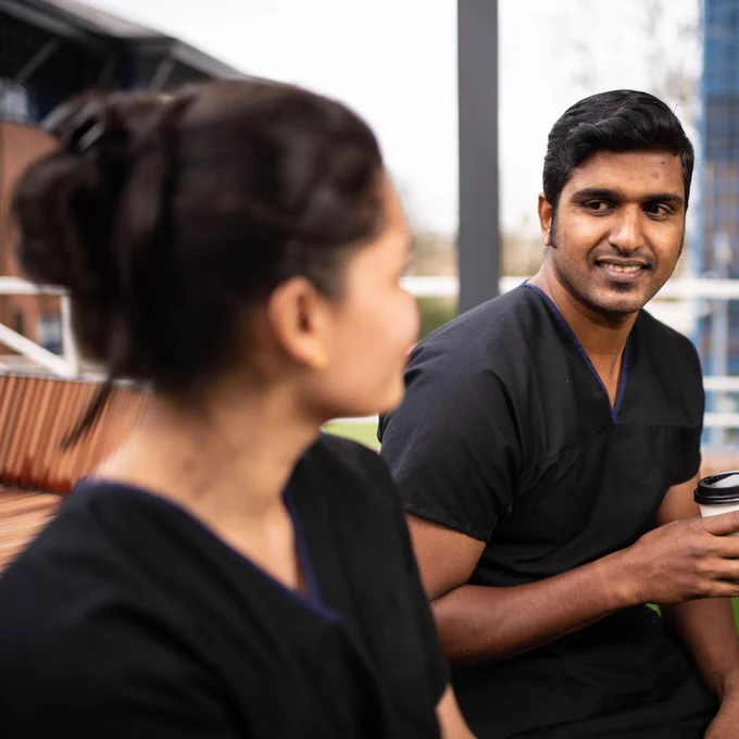 Two medical professionals in black scrubs are outdoors. One, facing away from the camera, is visible from the shoulder up. The other, holding a white coffee cup, looks to the right and smiles. They are seated on a wooden bench.