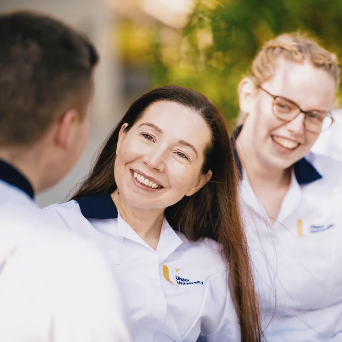 Three medical students in white uniforms with navy blue accents and the "Ulster University" logo are smiling and laughing outdoors. The woman in the foreground has long dark hair and is looking at someone off-camera to her left. Behind her, a woman with blonde hair and glasses smiles. 