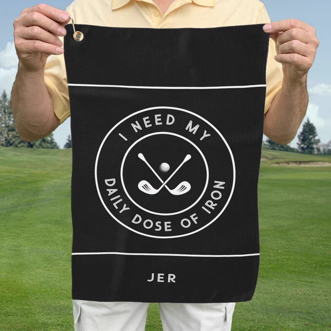 Black golf towel featuring crossed golf irons with a golf ball and bold 'I NEED MY DAILY DOSE OF IRON!' text in modern white font. Includes customizable monogram space for initials or name. All colors in this design may be customized.