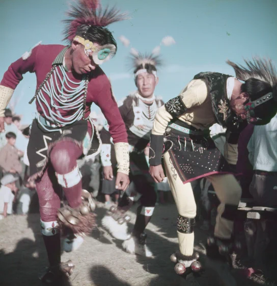 The photo shows two individuals performing a traditional dance, wearing regalia with feathers, beads, and bells. One dancer is in a red and black outfit with a raised leg, while the other, in yellow and black, is bending forward. A third person in similar attire is in the background, with a crowd watching. The scene appears to be outdoors during a cultural event.
