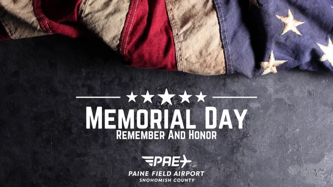 An American flag rests on dark slate stone. The words Memorial Day Remember and Honor appear in white below the flag with the official Paine Field logo.