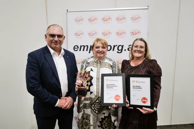 Two women stand together with a man, they're holding a trophy and certificates.