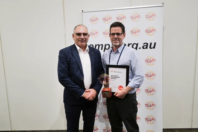 Two men stand together, one holds a trophy and a certificate.