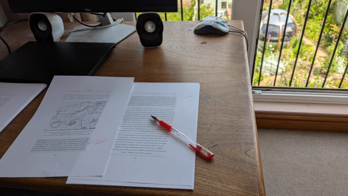 An open manuscript with red pen alterations on a traditional wooden desk overlooking a colourful garden