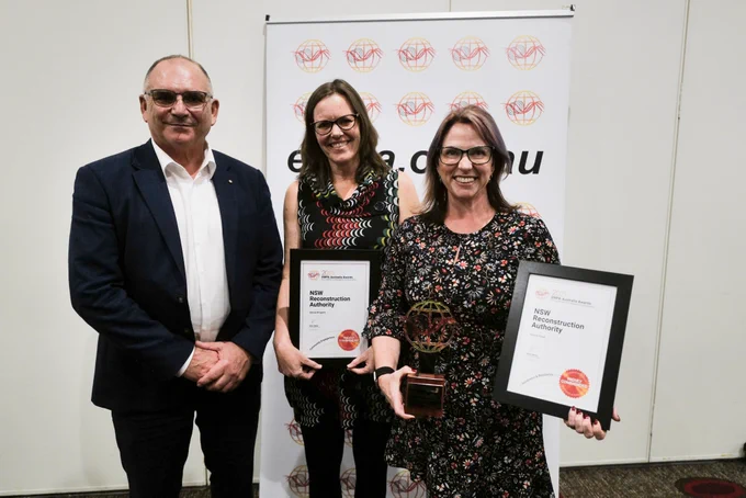 Two women stand together with a man, they're holding a trophy and certificates.