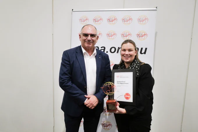 A man and a woman stand together, she's holding a trophy and a framed certificate.