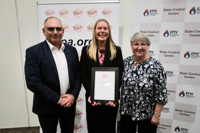 A woman with blonde hair holds a framed certificate. She stands between a man and a woman.