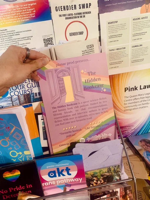 A POV shot of a hand tucking a lavender flyer decorated with rainbows and sparkles in amongst other similar flyers. The flyer is titled ‘The Hidden Bookcase.’