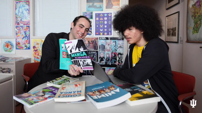Two individuals at a table, surrounded by various manga books, smiling and discussing a manga titled "Manga without Boundaries." The room is decorated with posters of anime and manga characters.
