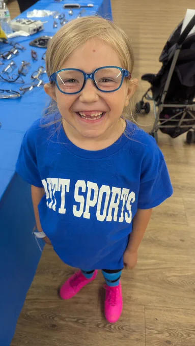 A young white girl with blond hair smiles at the camera. She's got blue glasses that match her blue T-shirt.