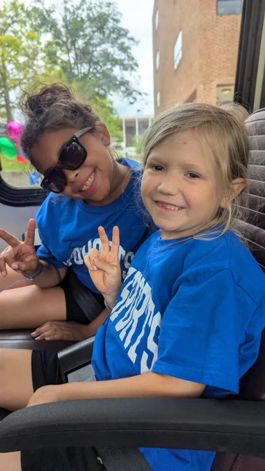 Two girls smile and make peace signs. One is Black with her hair in a bun and sunglasses. The other is white with her blond hair in a braid. They're sitting in a shuttle.