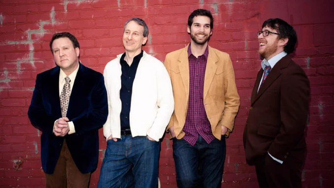 Four members of a band posing against a brick wall, smiling, dressed in semi-formal attire.