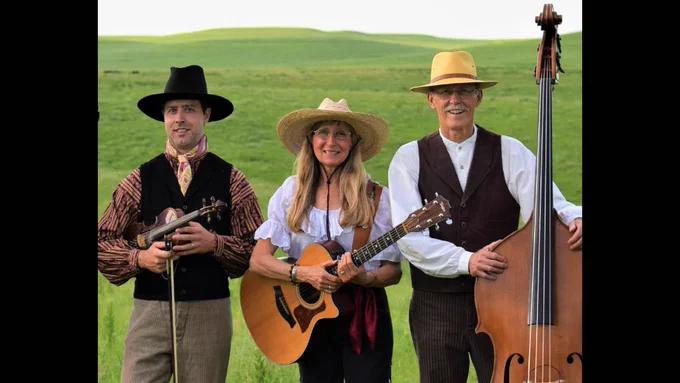 Three folk musicians in traditional attire posing with their instruments, a violin, a guitar, and a double bass, in a grassy field.
