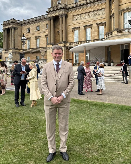A person in a beige suit standing in front of Buckingham Palace during an event, with guests in the background.