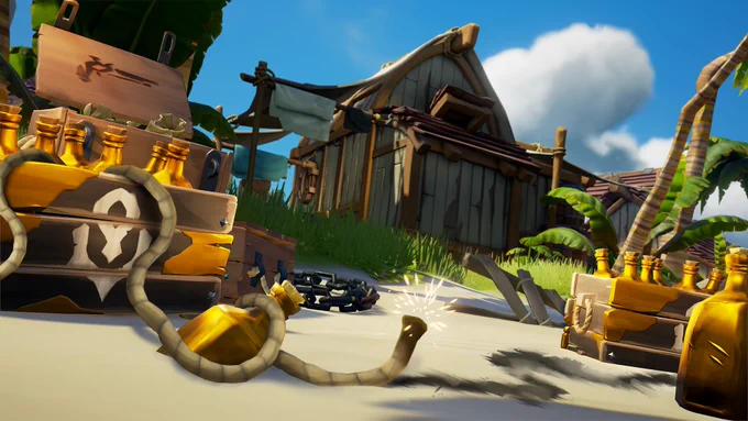 Multiple crates of golden bottles sit on a beach alongside an ammo box. The crates appear to have oddly-cut gold elements attached to some of the wooden planks and one is showing a white logo comprised of a large diamond with a much smaller diamond attached to its bottom tip, and a half oval circling underneath with a spike-like protrusion at its lowest point. A fuse runs from right to left, its right side fizzing and the left disappearing off-screen.