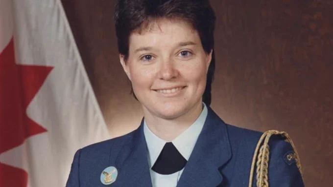 The photo shows Michelle Douglas, a woman dressed in a Canadian military uniform. She is wearing a blue jacket with a white shirt and a black tie, along with a shoulder cord and a badge featuring a maple leaf. The background includes the Canadian flag, indicating her association with the Canadian armed forces.