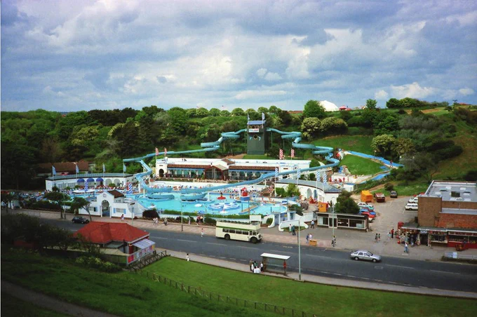 A vibrant 1980s scene of Water Splash World in Scarborough, UK, showing multiple blue water slides, a central outdoor pool area with people, and a bus parked on the road in front of the entrance.