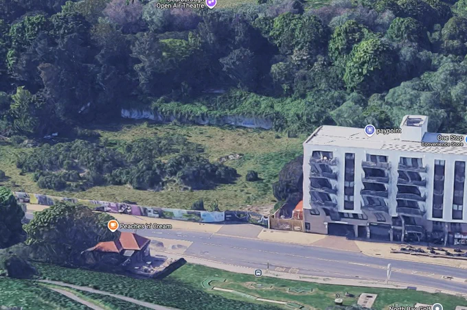 A modern aerial view of the former Water Splash World site in Scarborough, UK, now an overgrown grassy area behind a white apartment building and convenience store, with the Peaches 'n' Cream café still visible near the road.