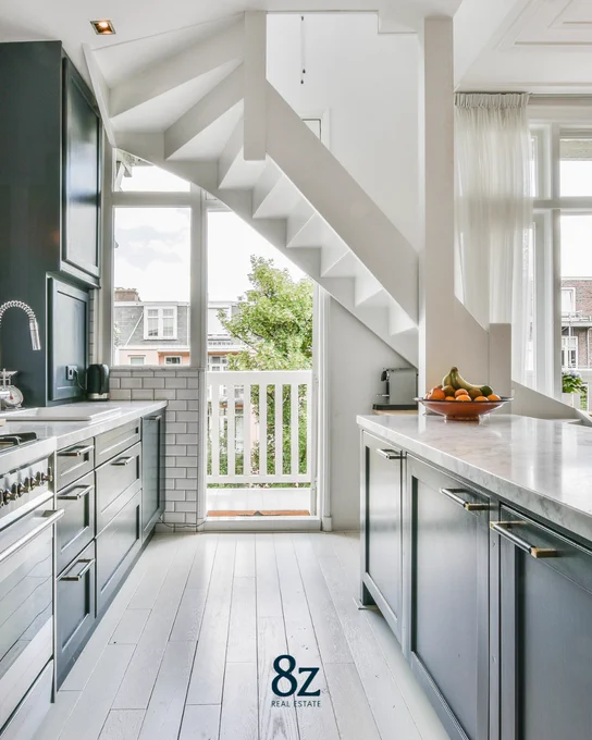 A modern kitchen featuring stainless steel appliances, grey cabinetry, and white brick-style backsplash. A staircase with a white railing ascends on the left, and natural light streams in through large windows with sheer curtains. A logo of "8z Real Estate" is visible in the foreground.