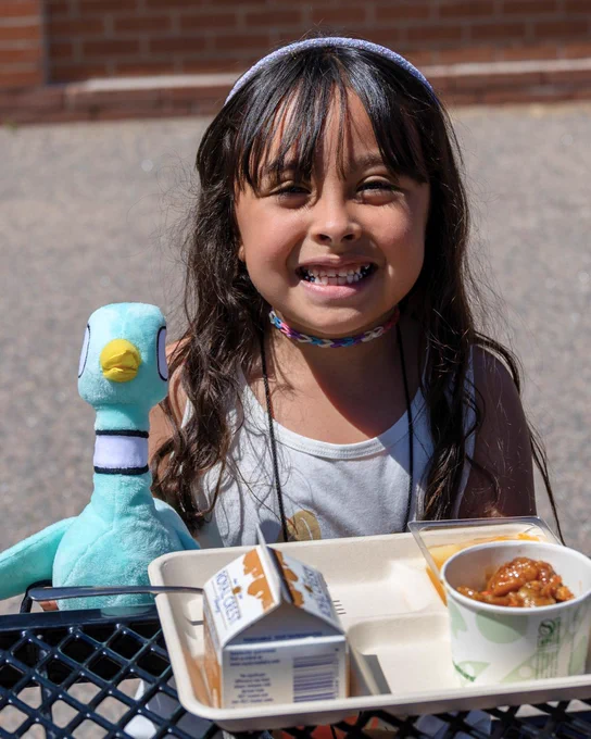 A child smiles holding a tray with food and a plush Pigeon toy, outdoors on a sunny day.