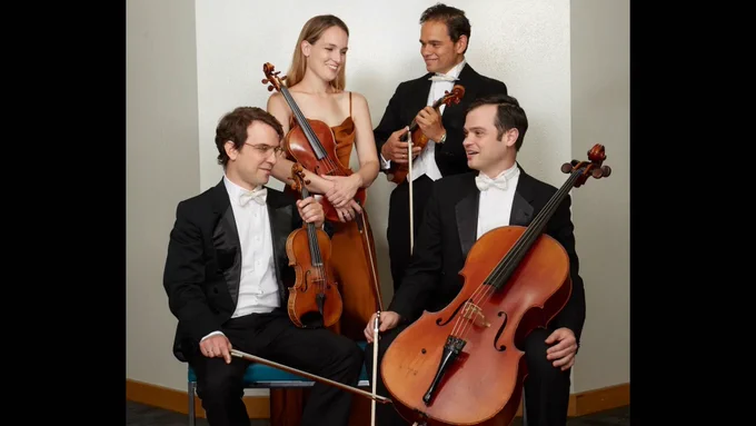 Four members of a classical string quartet smiling, holding their instruments: two violins, a viola, and a cello. They are dressed in formal attire.
