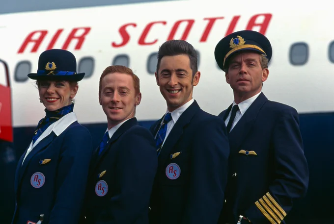 Four people in airline uniforms stand in front of a plane that reads AIR SCOTIA, smiling at the camera. 