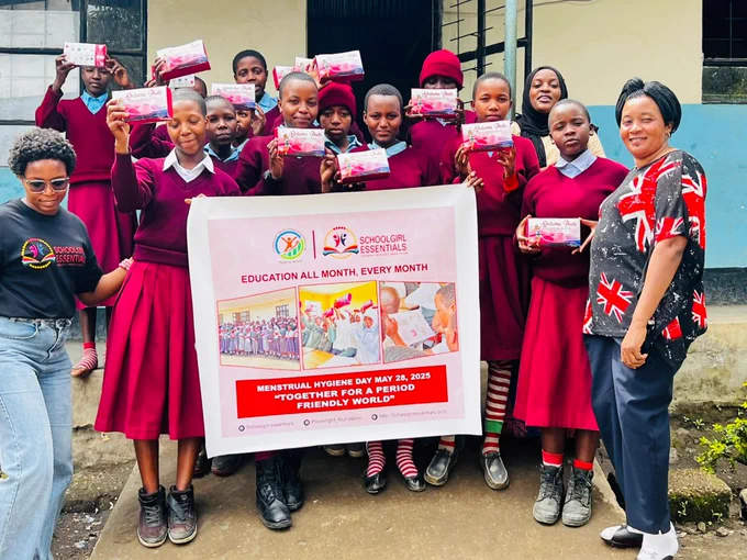 A group of around 12 Tanzania girls in burgundy school uniforms, alongside three women. The girls are holding an Education All Month, Every Month banner for World Menstrual Hygiene Day.