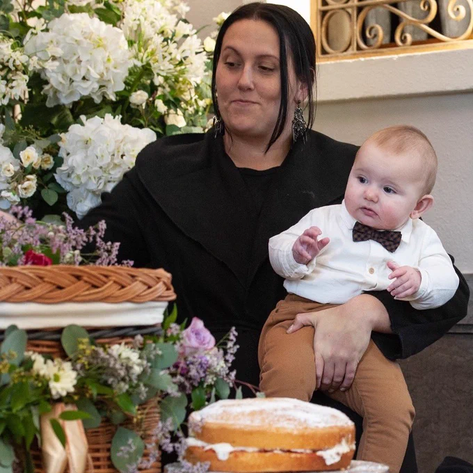 A dark haired woman dressed in black holds a baby dressed in a white shirt and bow tie. They are both looking at a willow coffin decorated with lilac coloured wildflowers. In front of the coffin is a Victoria Sponge cake filled with jam and cream.