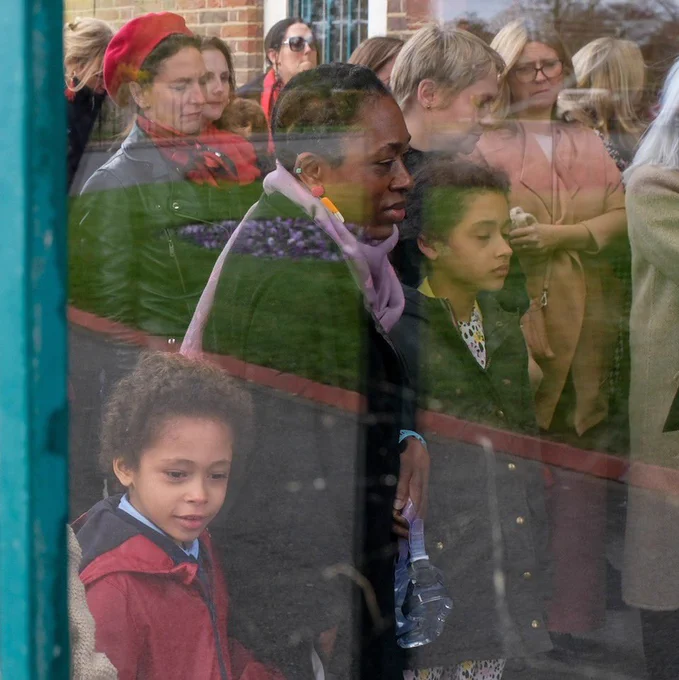 Two children stand among a group of adults, seen through a window