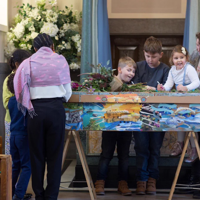 Two groups of children gather closely around a brightly decorated cardboard coffin with pens in their hands.