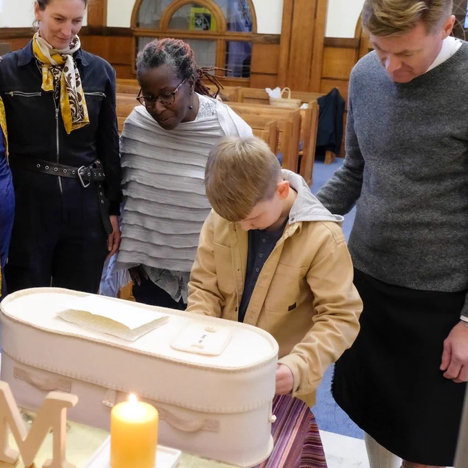 A fair haired young boy reaches out towards a small cream woollen coffin in front of a lit candle. Three adults stand by supportively.