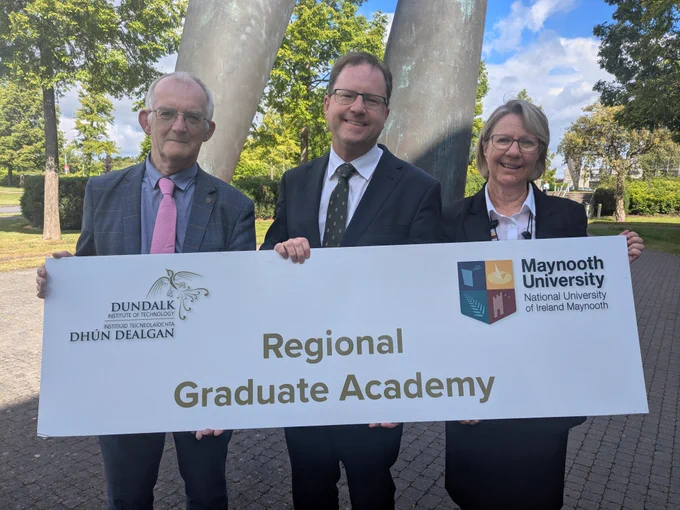 Three people stand outdoors holding a sign that reads "Regional Graduate Academy" with logos for Dundalk Institute of Technology and Maynooth University. They are dressed in formal attire, standing in front of a tree-filled background under a bright blue sky.