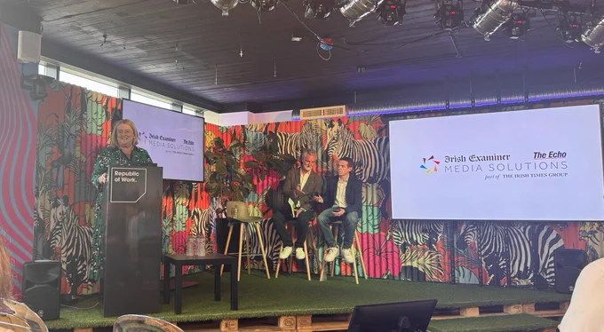 Woman speaking at podium with two male guest speakers and big screen in background. Screen shows Irish Examiner and The Echo logos.