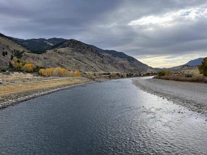 Photo of Yellowstone River in Montana. On a mostly overcast day, some sunlight reflects off the dark blue river. Mountains are seen in the background with yellow-brown vegetation at the base. Credit: NPS