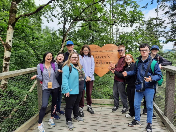 A group of TCPID students standing on a wooden platform in a forest with a wooden sign in the shape of a love heart. On the sign it says Lover's Lookout. 