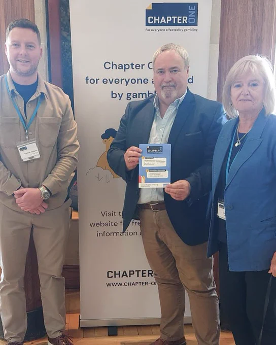 Pictured is Colm Gildernew MLA standing with reps of Chapter and gambling With Lives at Stormont.

In the backdrop a canvas can be seen with Chapter on it and information about their campaign to tackle gambling- the canvas is white with black wiring. Colm Gildernew MLA is holding a leaflet. A man standing to his right and a lady to his left. The herringbone floor can be seen.