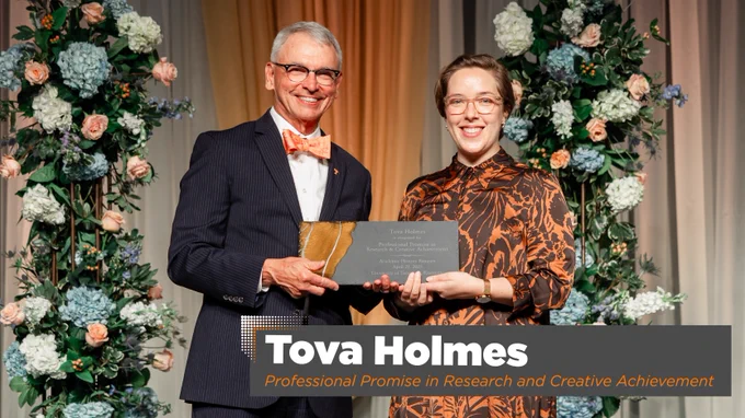Two individuals on stage at an awards ceremony, one presenting an award to the other named Tova Holmes for Professional Promise in Research and Creative Achievement, with a backdrop of flowers.