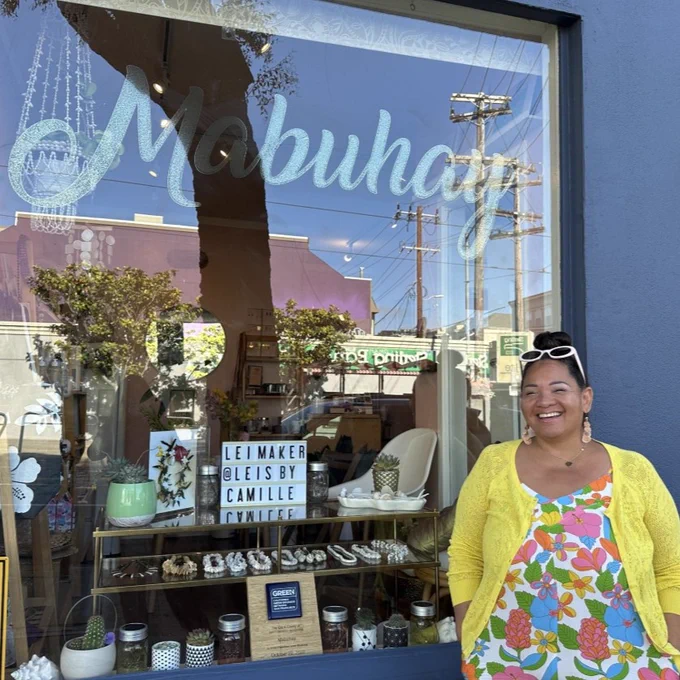 A person standing in front of the Mabuhay Beauty Boutique window, which displays various decorative items and signs mentioning "Lei Maker @LeisbyCamille Fontaine." This is a certified Green Business located in a sunny San Francisco area.