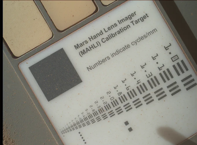 Close-up of the Mars Hand Lens Imager (MAHLI) Calibration Target on the Curiosity rover. The target features a dark gray square with a rough texture, several tan and beige color swatches, and a white panel with black resolution patterns labeled with numbers. Fine reddish Martian dust lightly coats the surface, giving it a speckled appearance.